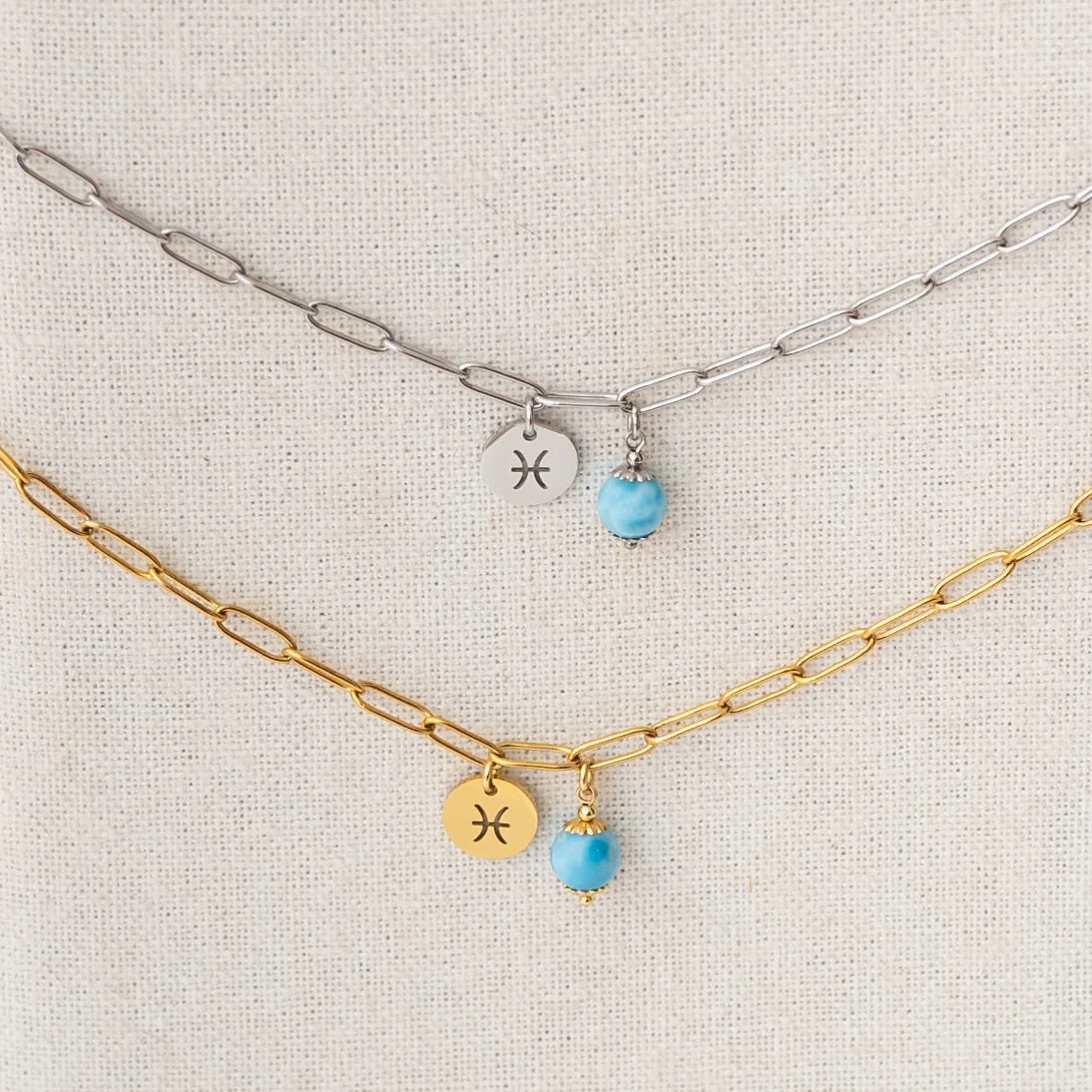 Deux colliers avec une perle de larimar de 6mm et un signe astrologique suspendu à un présentoir, en vue proche. Colliers en acier inoxydable 316L argent et doré. Larimar origine République Dominicaine.