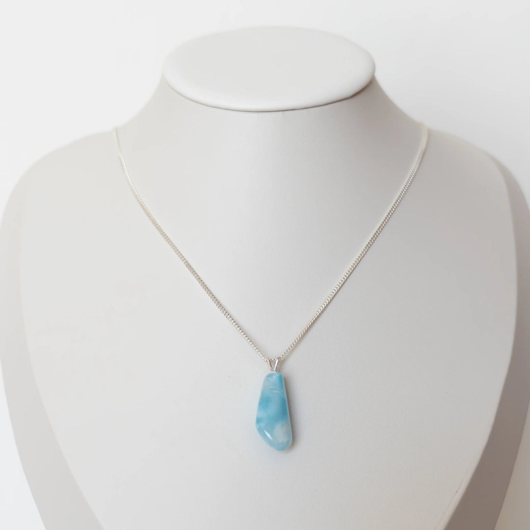 Pierre de Larimar de forme triangulaire montée sur une chaîne en argent 925 et posée sur un présentoir en forme de buste. Le Larimar est une pierre de couleur bleue unique au monde.