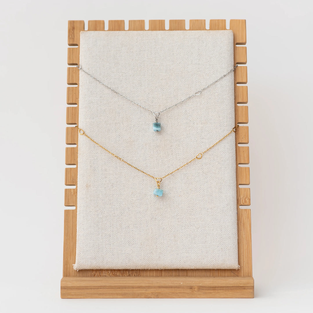 Un collier en acier inoxydable argenté et un collier en acier inoxydable 316L doré avec chacun un pendentif en Larimar bleu, sont exposé sur un présentoir à collier en bois sur fond blanc.