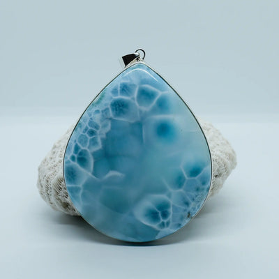 Pendentif en argent 925 ornant une magnifique pierre naturelle de Larimar de forme ovale. Cette pierre de Larimar à plusieurs nuances de bleu, elle est posée contre un coquillage.