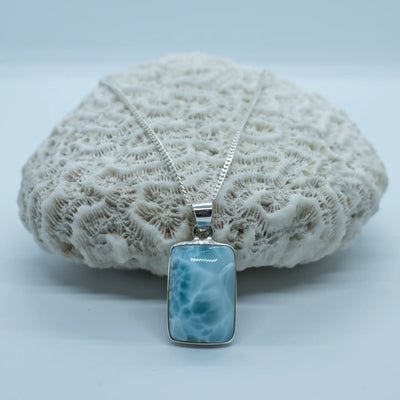 Pierre naturelle de larimar de couleur bleu avec des parties de différentes couleurs de bleu, faite en larimar, posée devant un coquillage blanc de type corail.