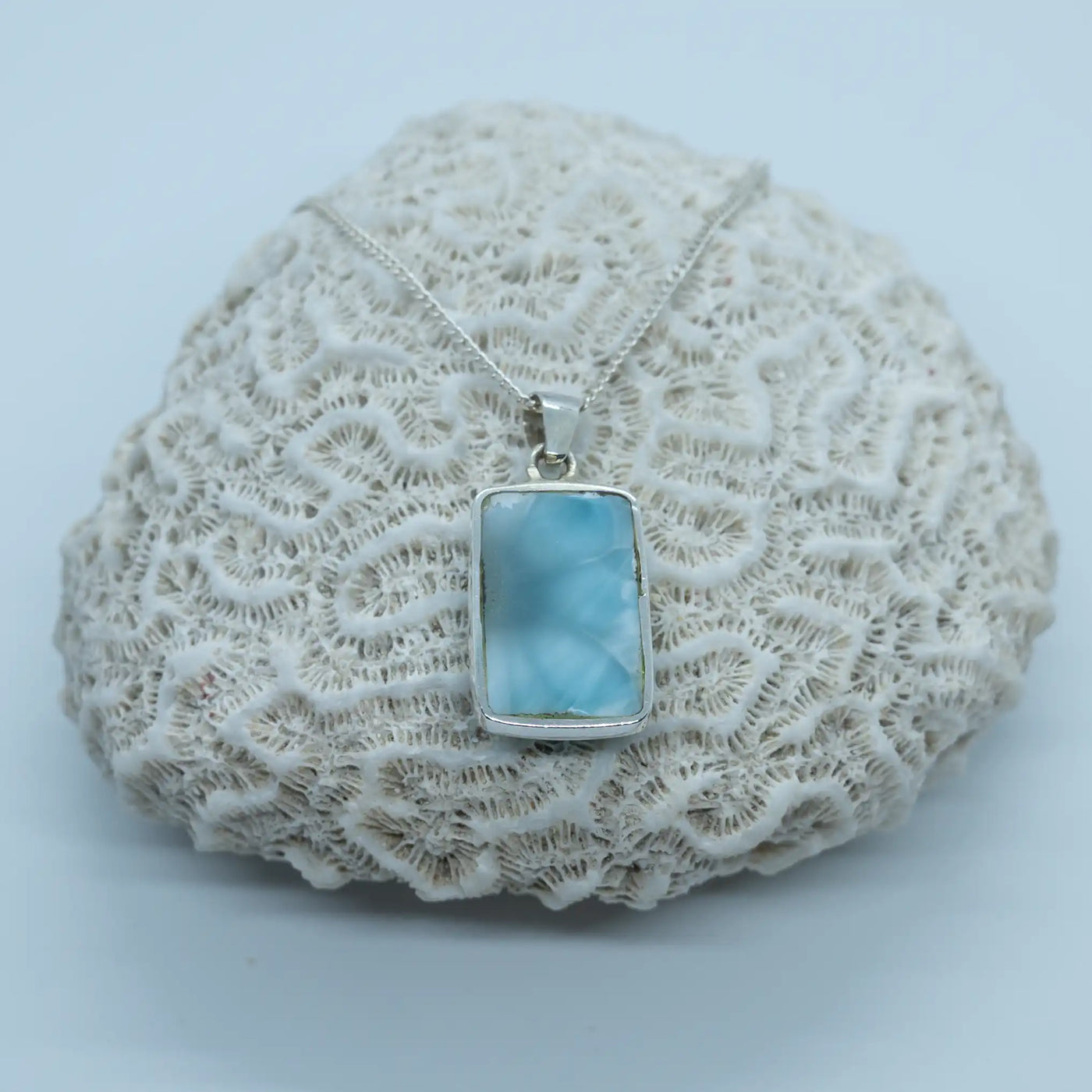 Cabochon rectangulaire bleu, en Larimar de République Dominicaine, posé sur un coquillage. Seul la face verso du cabochon est visible, elle est non serti pour avoir la pierre naturelle contre la peau.