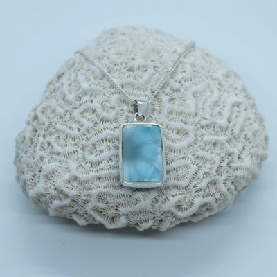 Cabochon rectangulaire bleu, en Larimar de République Dominicaine, posé sur un coquillage. Seul la face verso du cabochon est visible, elle est non serti pour avoir la pierre naturelle contre la peau.