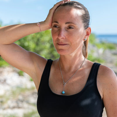 Femme accoudé à une brache d'arbre, avec la mer et les rochers en arrière plan, portant un collier en acier inoxydable 316L argenté, s'ouvrant et se fermant sur le devant via un fermoir en T sur lequel est attaché une perle de larimar de 11mm de diamètre au bout d'un petit bout de chaîne. 