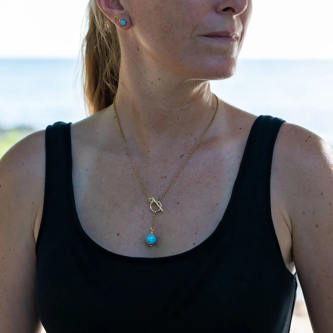 Femme vue de face portant un collier couleur or en acier inoxydable 316L. Collier avec fermoir en T sur le devant, avec une perle de larimar bleue pendante.