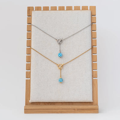 Présentoir avec deux colliers en acier inoxydable 316L, l'un est argenté, l'autre est doré. Aussi appelé collier T de part son système de fermeture avec un anneau dans lequel vient se loger une pièce en forme de T. Une perle de larimar de 11mm le long d'une chaine de 2cm est raccroché à cette anneau fermoir.