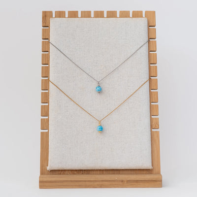 Présentoir avec un collier en acier inoxydable 316L argenté et un collier en acier inoxydable 316L doré. Chaque collier possède une perle de larimar en pendant.