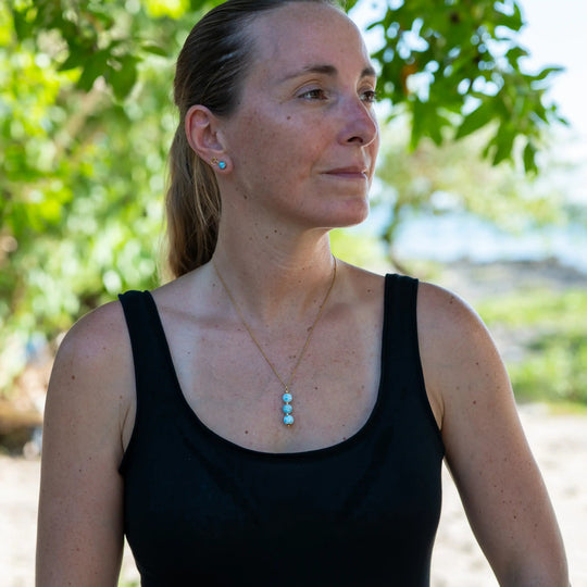Femme prise en photo de face avec un arrière plan flou représentant une cote rocheuse de bord de mer, portant un collier avec trois perles de larimar bleues. Collier de couleur doré en acier inoxydable.