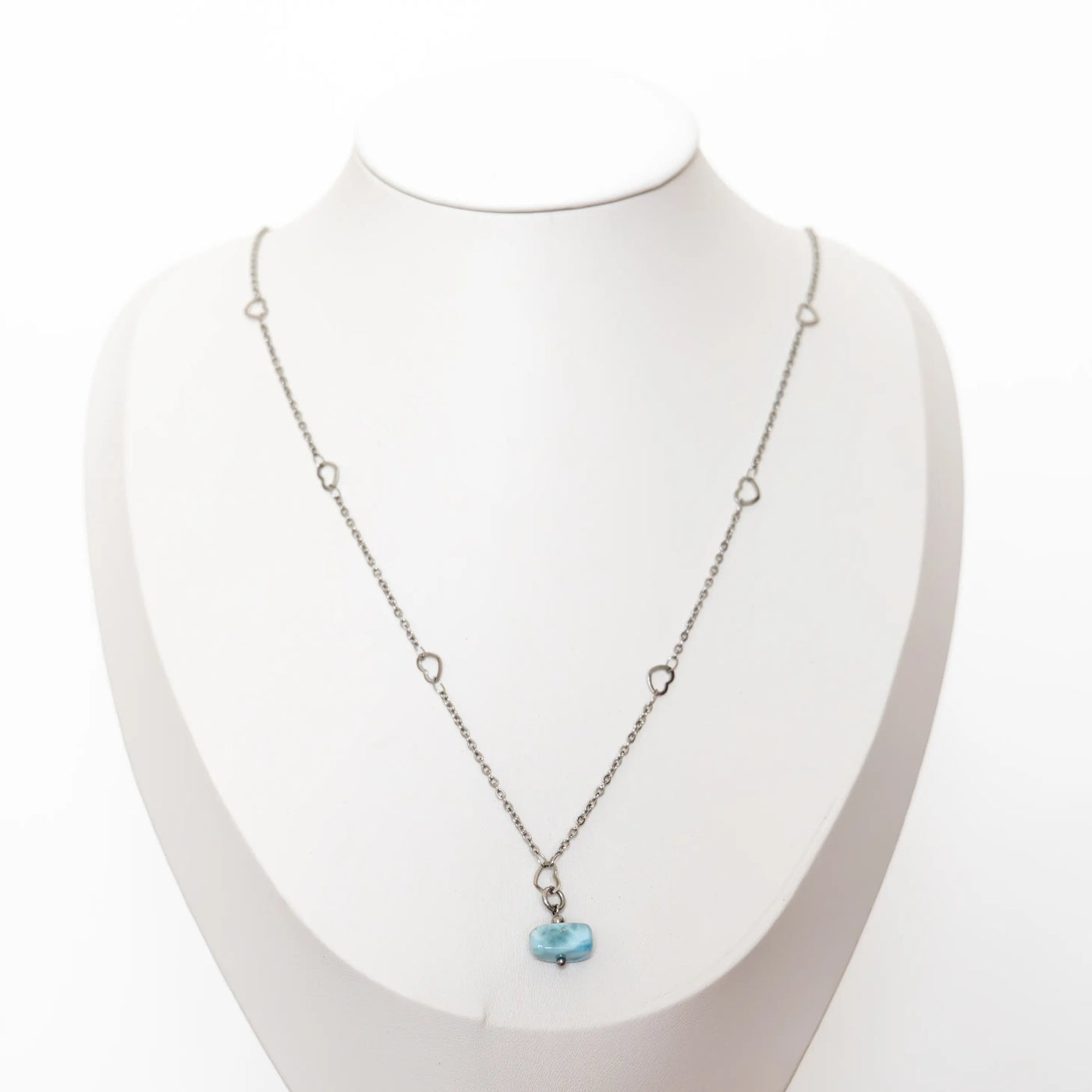 Collier avec une chaîne en acier inoxydable 316 de couleur argentée posé sur un présentoir en forme de buste. Ce collier à un pendentif en pierre de Larimar de République Dominicaine. La chaîne possède plusieurs petits cœurs. Bijou fait à la main.