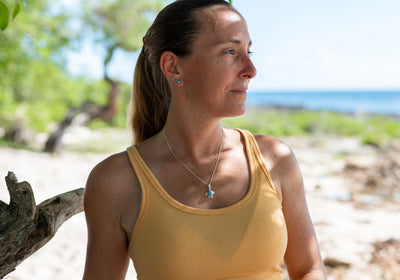 Femme portant un collier en forme de tortue avec un cabochon de Larimar en son centre. Le collier est fait en argent 925,la femme porte un débardeur jaune, elle se trouve sur une plage que l'on voit flou en arrière plan.