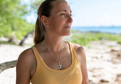 Femme se tenant devant une plage, portant un débardeur jaune ainsi qu'un collier fait en argent 925 avec un pendentif en forme de tortue avec un cabochon en Larimar.