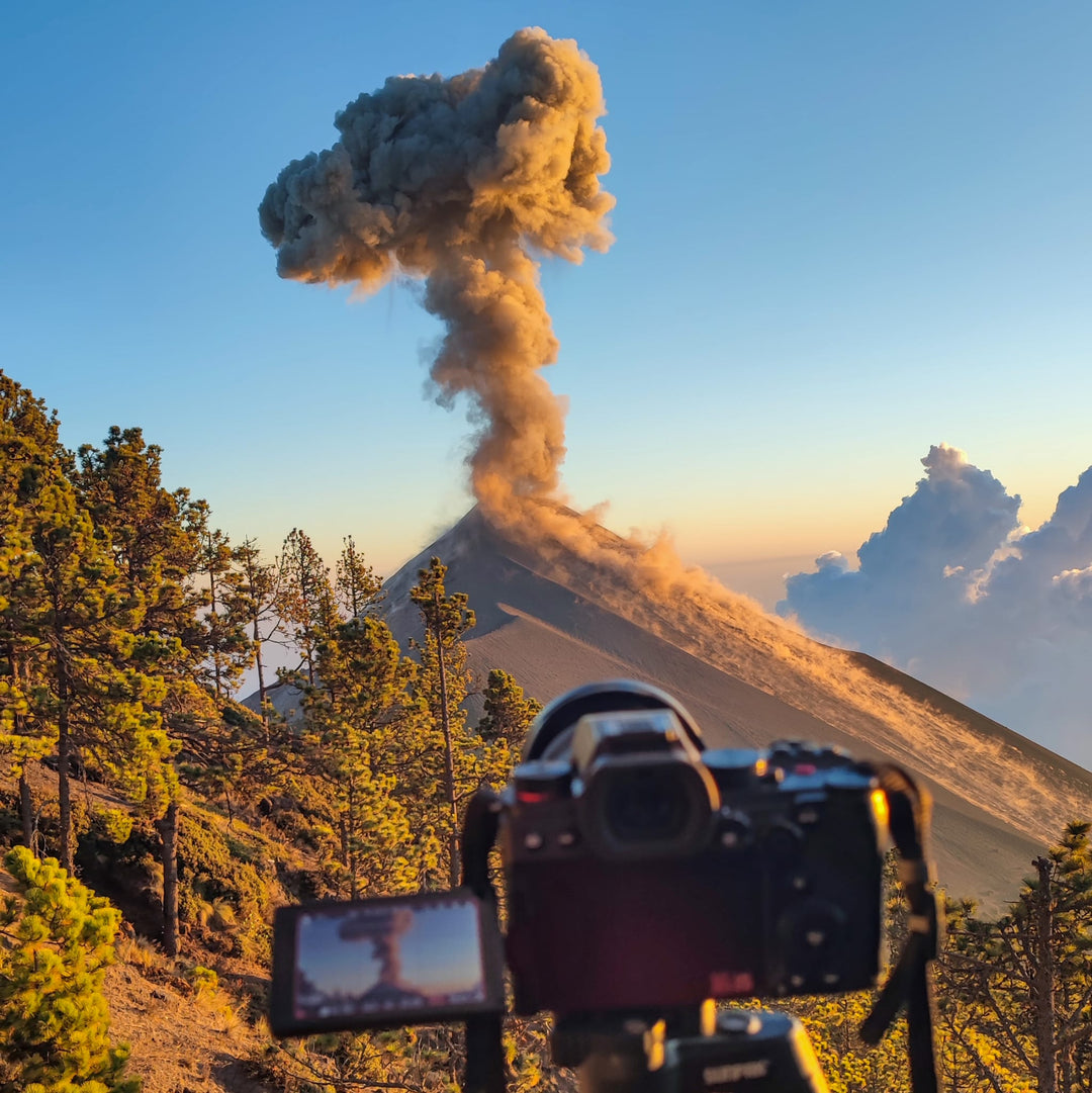 Photo du volcan El Fuego pris depuis le point de vue en haut du volcan Acatenango. On aperçoit en premier plan un appareil photo en train de réaliser un time lapse et le volcan El fuego en fond en train de cracher un panache de fumée.