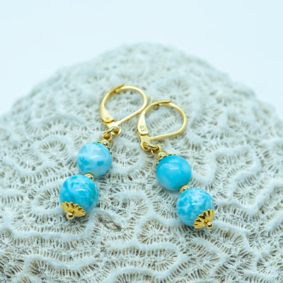 Paire de boucles d'oreilles dorées posées sur un coquillage sur fond blanc. Boucles d'oreilles en acier inoxydable 316L avec deux perles de larimar origine République Dominicaine en pendants.
