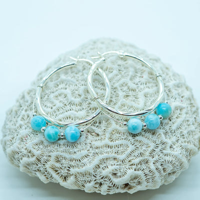 Trois perles de larimar de République Dominicaine montées sur une paire de boucles d'oreilles créoles en argent 925. Les boucles d'oreilles sont posées sur un coquillage.