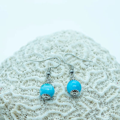 Deux boucles d'oreilles argentées posées sur un coquillage, faite en acier inoxydable 316L. Chaque boucle à une perle de larimar de République Dominicaine en pendant.