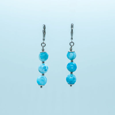 Photo d'une paire de boucles d'oreilles vue de face, faite en acier inoxydable 316L argenté. Chaque boucle à un pendant avec trois perles de larimar.