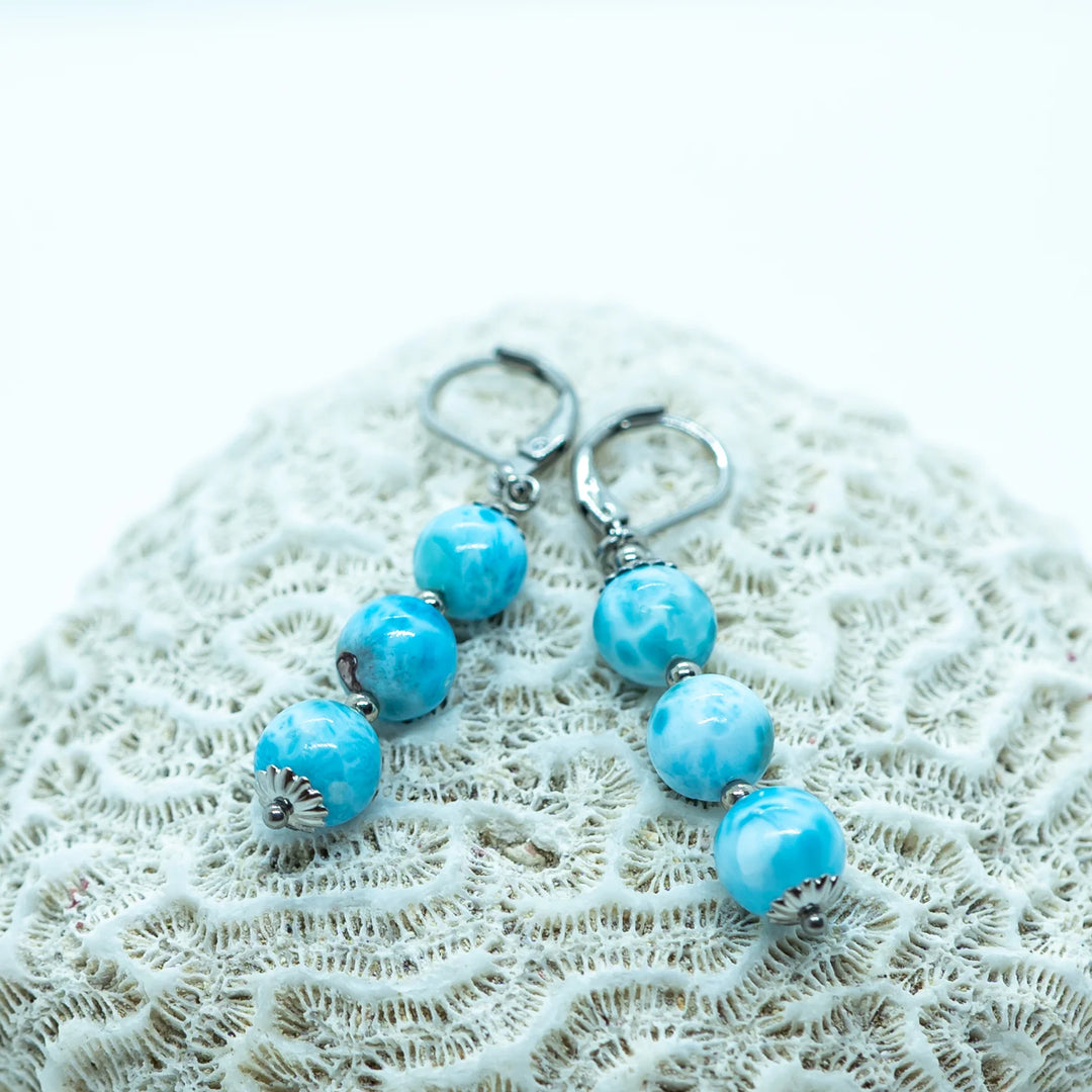 Deux boucles d'oreilles identiques, de couleur argentée, faite en acier inoxydable 316L avec trois perles de larimar en pendant, posées sur un coquillage.