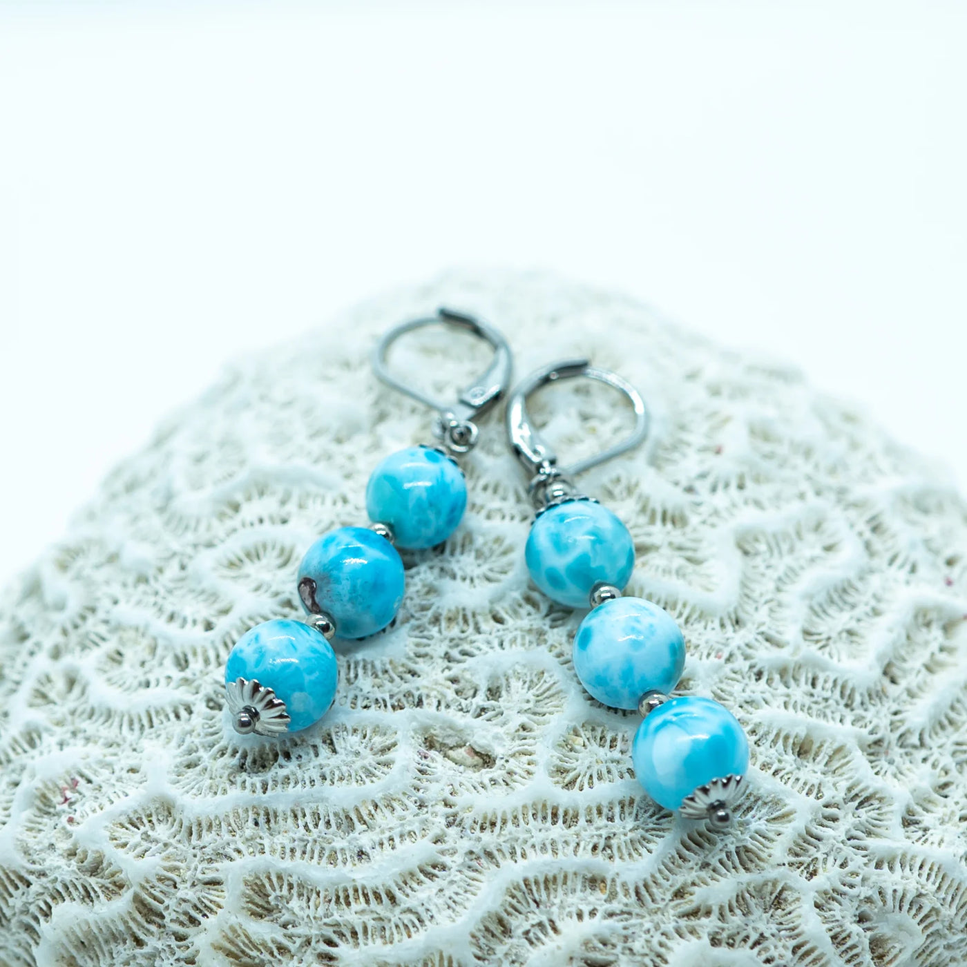 Deux boucles d'oreilles identiques, de couleur argentée, faite en acier inoxydable 316L avec trois perles de larimar en pendant, posées sur un coquillage.