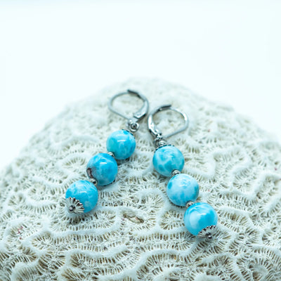 Deux boucles d'oreilles identiques, de couleur argentée, faite en acier inoxydable 316L avec trois perles de larimar en pendant, posées sur un coquillage.