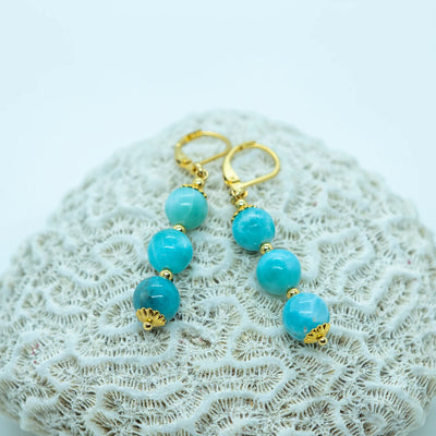 Paire de boucles d'oreilles dorées posées sur un coquillage vue de prés. Les boucles d'oreilles sont en acier inoxydable 316L et possèdent trois perles de larimar en pendant.