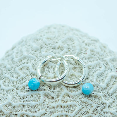 Boucles d'oreilles créoles en argent 925 posées sur un coquillage sur fond blanc. Perle de larimar bleue origine République Dominicaine en pendant, boucles d'oreilles texturées