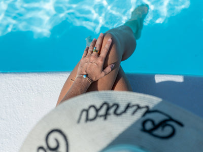Vue du dessus d'une femme assise sur le bord d'une piscine avec les jambes croisées et les pieds dans l'eau. Elle porte un bracelet en cordon doré avec une perle de larimar bleu au poignet gauche et à la main posée sur le genou.
