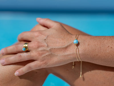 Bracelet en cordon doré avec une perle de larimar de couleur bleue, porté sur le poignet gauche d'une femme, assise sur le bord d'une piscine.