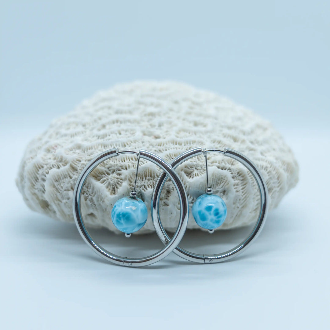 Magnifique perles en Larimar de République Dominicaine avec plusieurs nuances de bleu et un maillage blanc, montées sur une paire de créoles en acier inoxydable 316L et posées devant un coquillage.