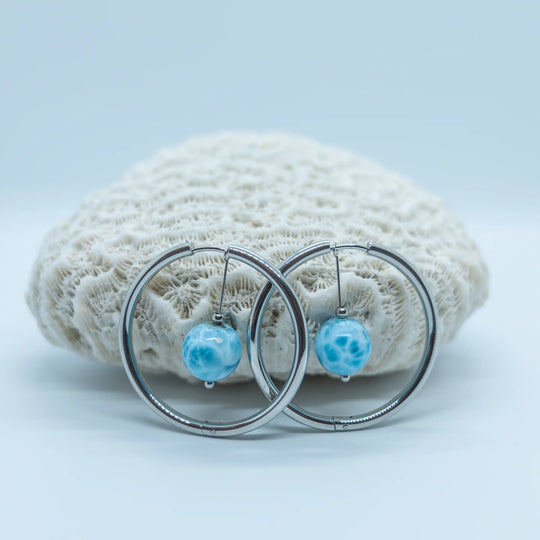 Magnifique perles en Larimar de République Dominicaine avec plusieurs nuances de bleu et un maillage blanc, montées sur une paire de créoles en acier inoxydable 316L et posées devant un coquillage.