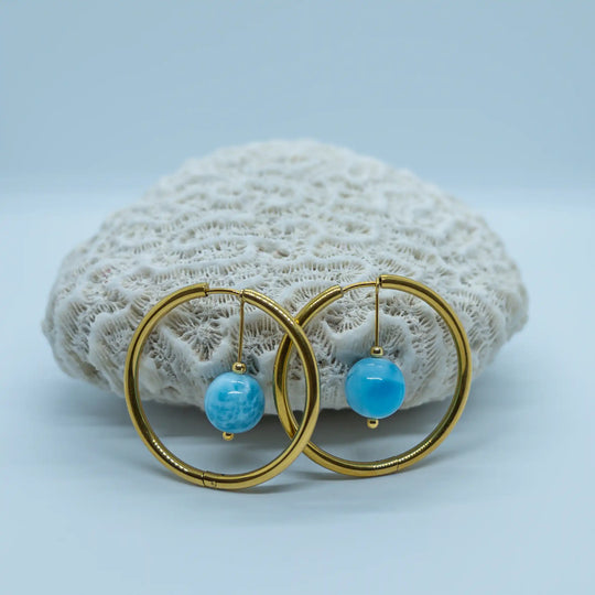 Créoles avec leur perle de Larimar bleu posées devant un coquillage blanc sur fond bleu-gris. Bijou unique, fait à la main en République Dominicaine, les créoles sont en acier inoxydable 316L doré.