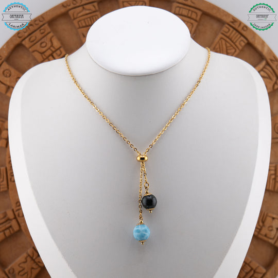 Deux perles naturelles, la première en Jade Jaguar et la deuxième en Larimar, sont montées sur un collier doré avec une bague en caoutchouc permettant de régler la position de chacune des perles.