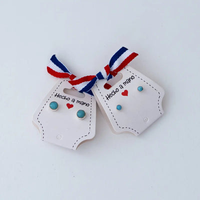 Photo de deux types de puces d'oreilles vue de face, monté sur un packaging avec un ruban bleu, blanc et rouge. Puces d'oreilles rondes avec un cabochon en larimar. Une des paires de puces et plus grosse que l'autre.