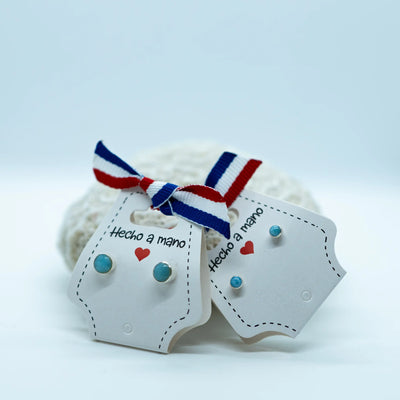Puces d'oreilles ronde avec une pierre de larimar sertie. Deux tailles de puces d'oreilles, chacune étant faite en argent 925. Les puces d'oreilles sont montées sur un packaging en carton blanc avec un ruban.