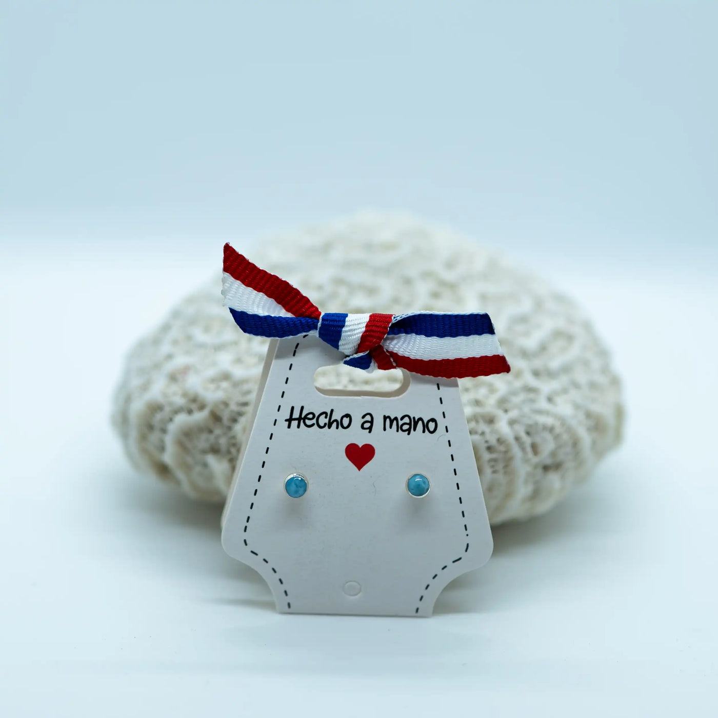 Paire de puces d'oreilles très fines avec une pierre de larimar. Faites en argent 925, elles sont montées sur un packaging en carton blanc avec un nœud de ruban bleu, blanc et rouge 