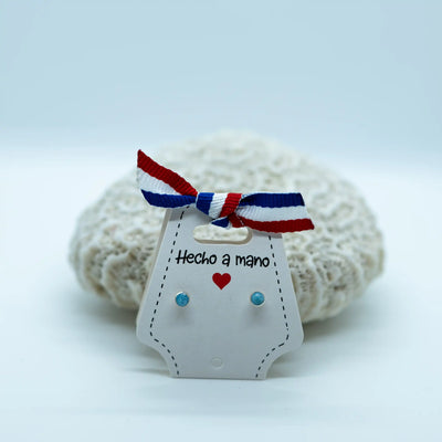 Paire de puces d'oreilles très fines avec une pierre de larimar. Faites en argent 925, elles sont montées sur un packaging en carton blanc avec un nœud de ruban bleu, blanc et rouge 