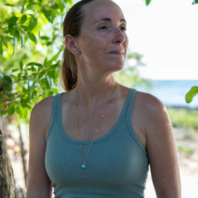 Sautoir doré en acier inoxydable 316L et son pendentif en Larimar, une pierre naturelle de République Dominicaine, porté par une femme avec un débardeur couleur kaki. L'arrière plan est flou et semble être une plage.
