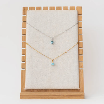 Présentoir en bois avec deux sautoirs en acier inoxydable 316L, l'un de couleur argenté et l'autre de couleur doré. Ces sautoirs ont un pendentif en Larimar bleu venant de République Dominicaine.