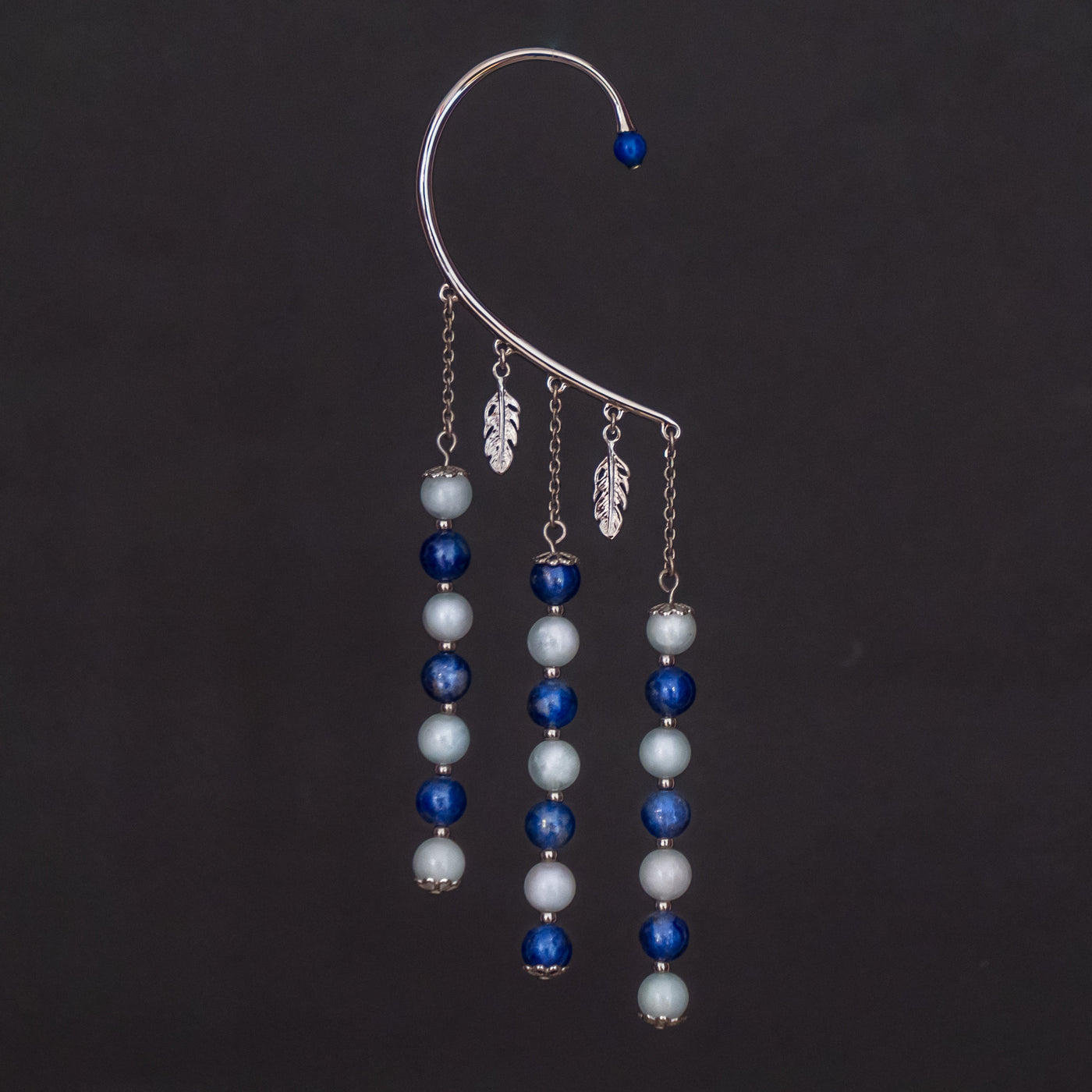 Tour d'oreille de couleur argenté avec ses deux breloque en forme de feuille, ses trois pendants avec des perles bleu de deux teintes différentes. Une perle d'énergie termine le tour d'oreille sur le haut.